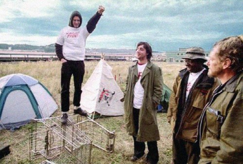 John Paul Cupp (c) in trench coat when he was homeless living in Portland Oregon in the early 2000’s when he became the chief U.S representative of the Pyongyang sanctioned group of U.S. supporters of North Korea. North Korean media heralded Cupp as a “prominent U.S. public figure.”